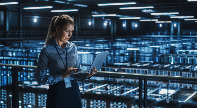A professional woman in a business suit poses in a large server room, surrounded by numerous servers and technology.