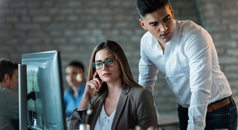 Two people in an office, one working at a computer while the other offers assistance