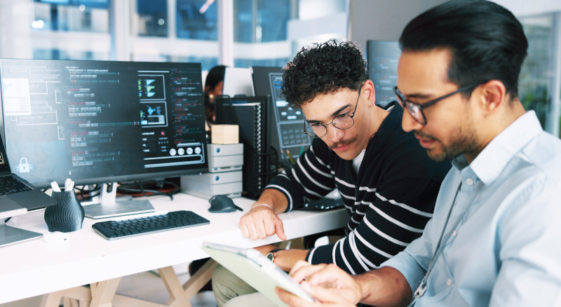 Two men examine a tablet computer while seated in front of a desktop computer.