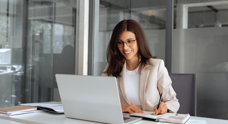Smiling woman in a beige blazer works on a laptop at a modern office desk. She writes in a notebook, conveying focus and professionalism.