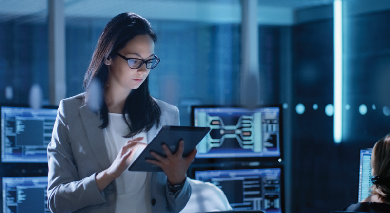 A focused professional in a modern office uses a tablet, surrounded by computer screens with data. The setting conveys a high-tech, analytical atmosphere.