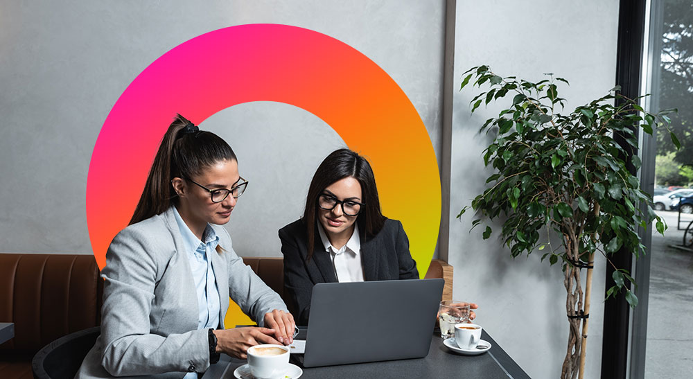 Two business women looking at laptop at a coffee shop with SHI logo behind them