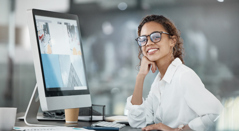 A smiling woman with glasses sits at a desk in an office, leaning on her hand. She faces a computer displaying images.