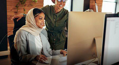 Two individuals in an office setting, one seated at a computer and the other standing behind, engaged in a discussion