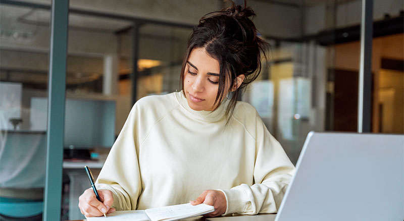  A woman sits at a desk, writing in a notebook with a focused expression