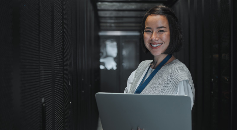 A person holding a laptop in a server room with rows of data racks on both sides