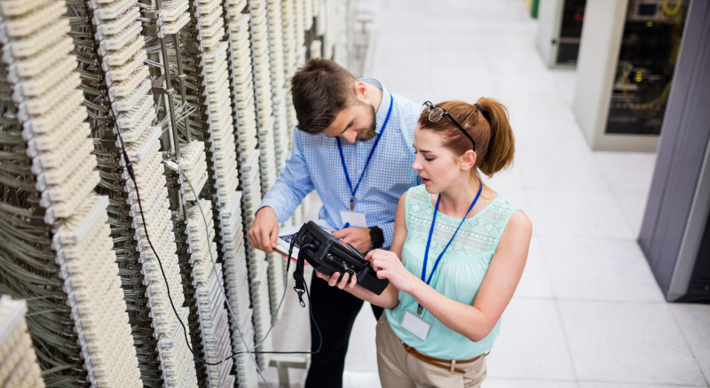 Two technicians examining a device in front of a server rack with cables and equipment