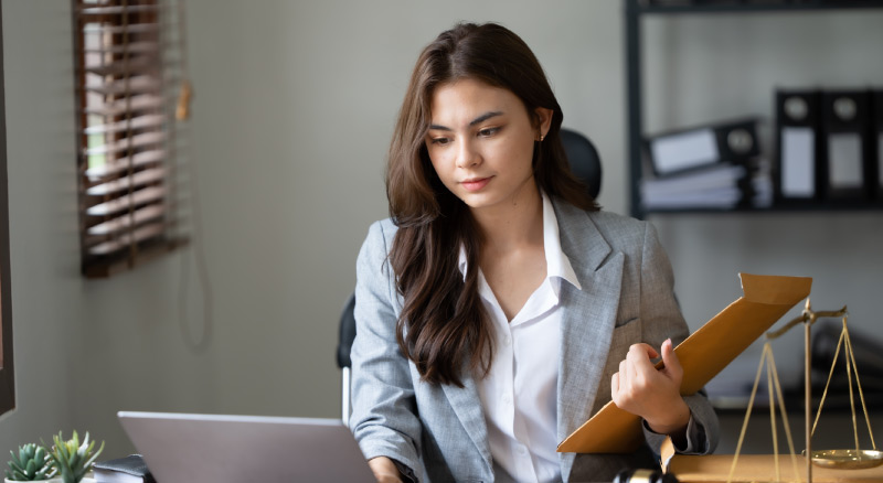 A professional individual holding a folder, with a laptop and a golden scale on the desk, in an office setting