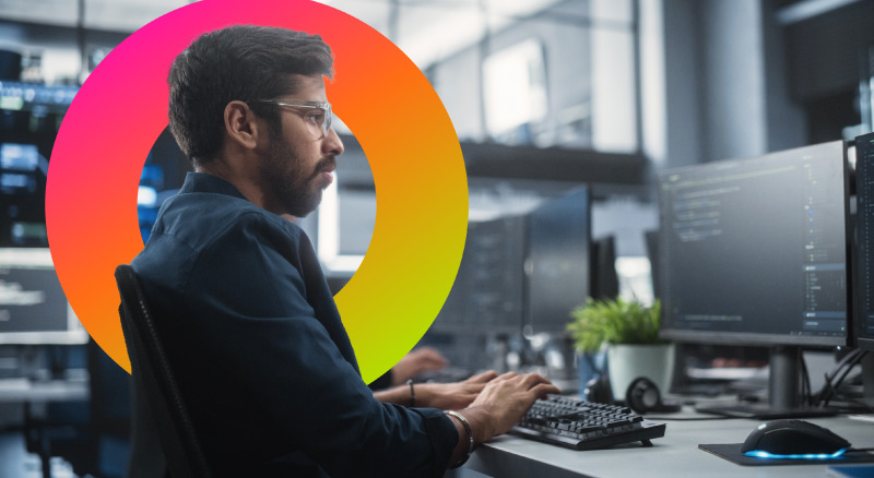 The person is sitting at a desk with two computer monitors, a keyboard, and a plant.