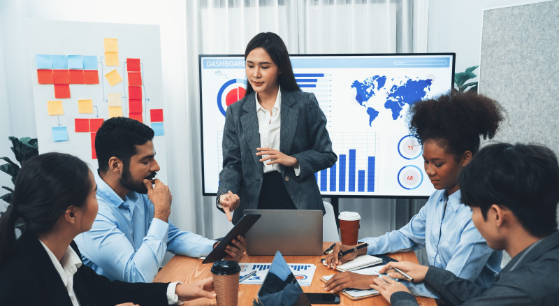 A group of professionals in a meeting room discussing data displayed on a screen.