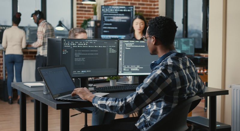 A person is working on a computer with multiple monitors in a busy office environment