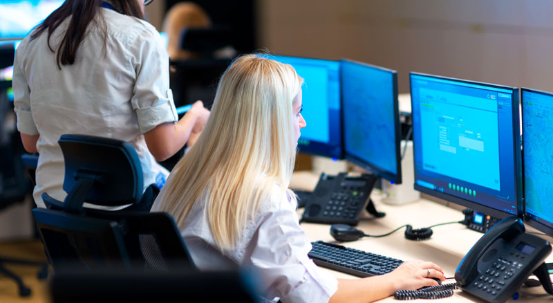 Two individuals working at a multi-monitor computer setup in a busy office environment