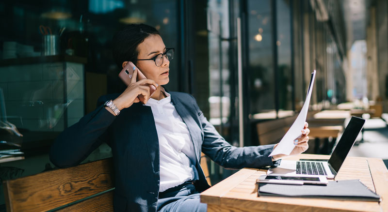 A person is seated at a wooden table, reviewing a document while also using a laptop and talking on the phone