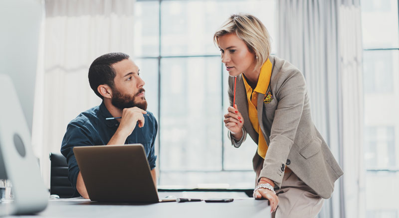 Two people in business attire having a conversation in a well-lit office behind a laptop