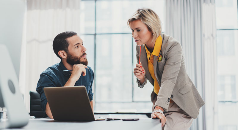 Two people in business attire having a conversation in a well-lit office behind a laptop