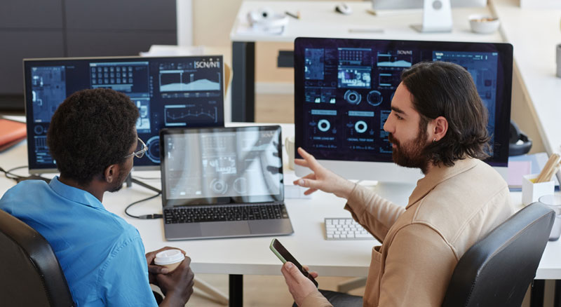 Two people in an office with multiple computer monitors displaying complex interfaces