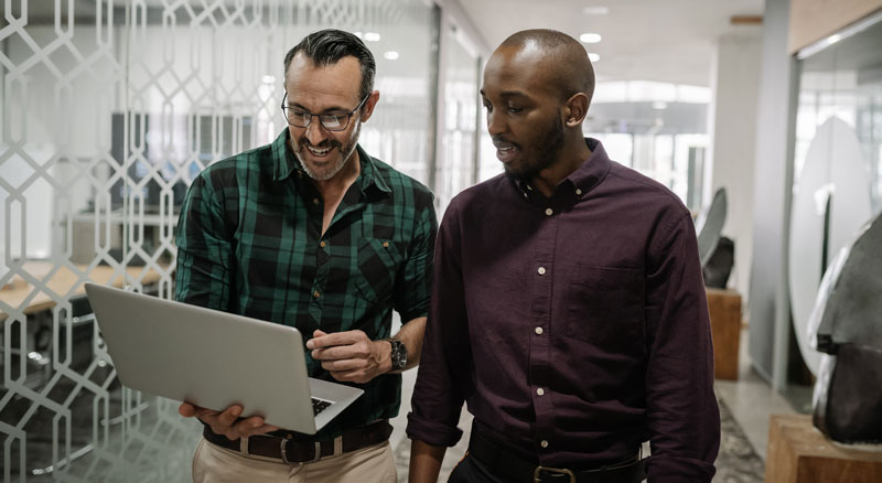 Two individuals in an office environment, walking and looking at a laptop one of them is holding