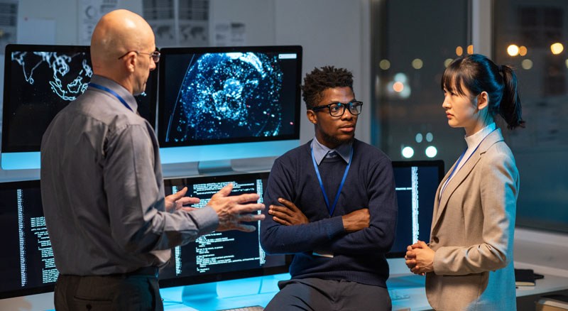 Three professionals discussing data displayed on multiple computer monitors