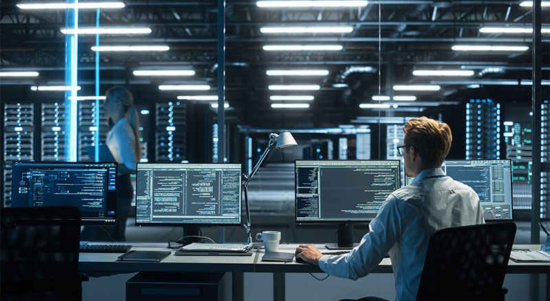 A man sitting at a desk in a large room with multiple computers, working diligently.