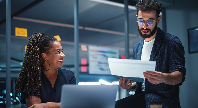 A man and woman sitting at a desk, working on a laptop together.