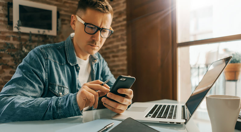 A person using a smartphone near an open laptop in a warm home office