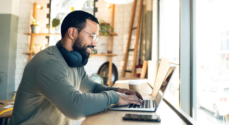 A person is working on a laptop with headphones on, at a wooden table beside a window, with a cup and smartphone nearby