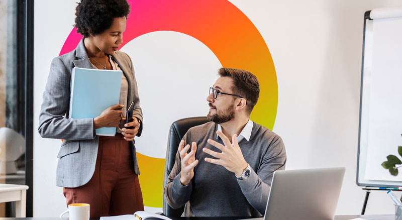 Two professionals in a modern office, one standing and holding a folder, the other seated with a laptop, engaged in a conversation