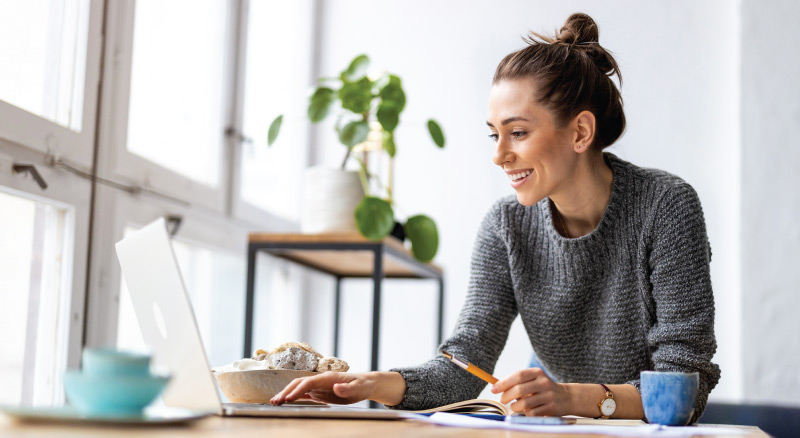 Person working on laptop with notebook and snack in bright room
