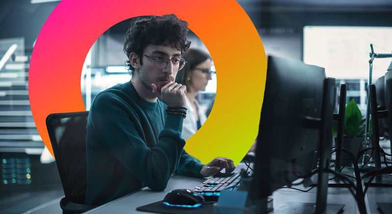 A person sits at a desk with multiple monitors and electronic devices, working on a computer