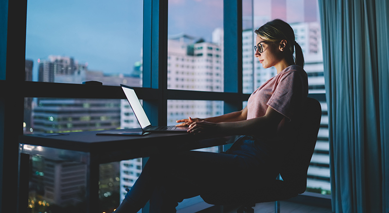 A person working on a laptop in a room with a view of the cityscape during the evening