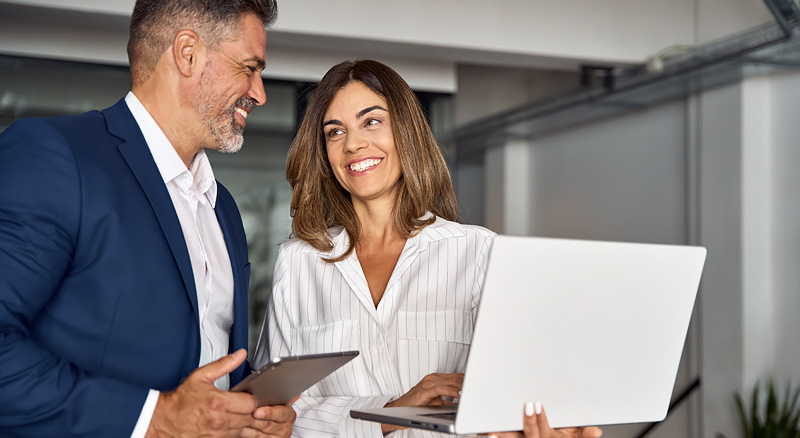 Two employees converse while holding a laptop and tablet