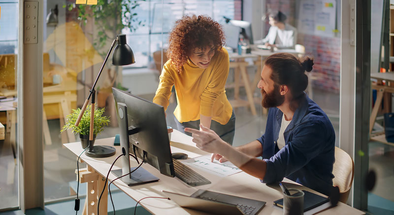 Two colleagues are engaged in a discussion at a well-organized workspace, with one person pointing towards the computer screen