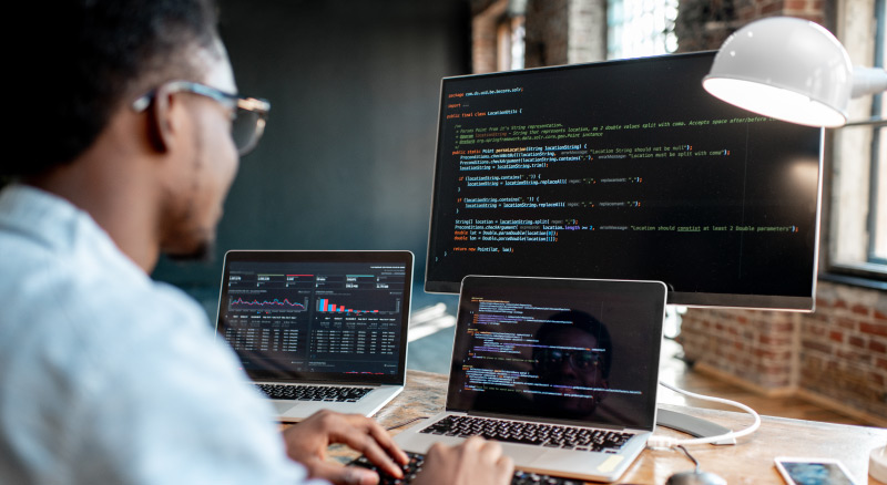 An individual is working at a desk with multiple computers, one of which displays lines of code, while the others show graphical data and charts