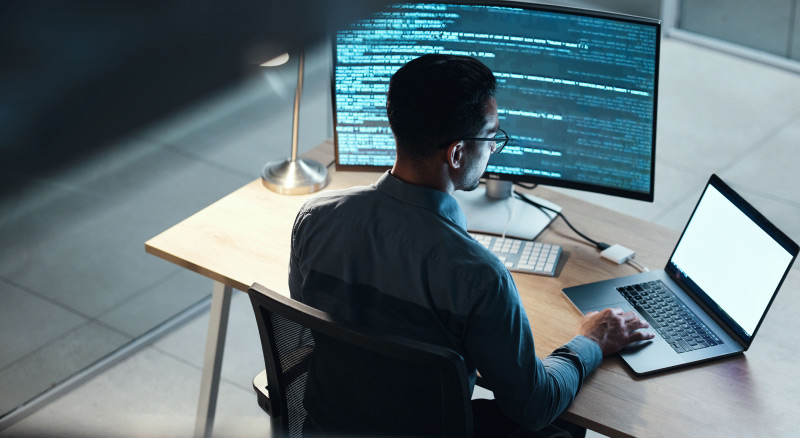 An individual is seated at a desk, working on a laptop with a large monitor displaying lines of code, in a well-lit room