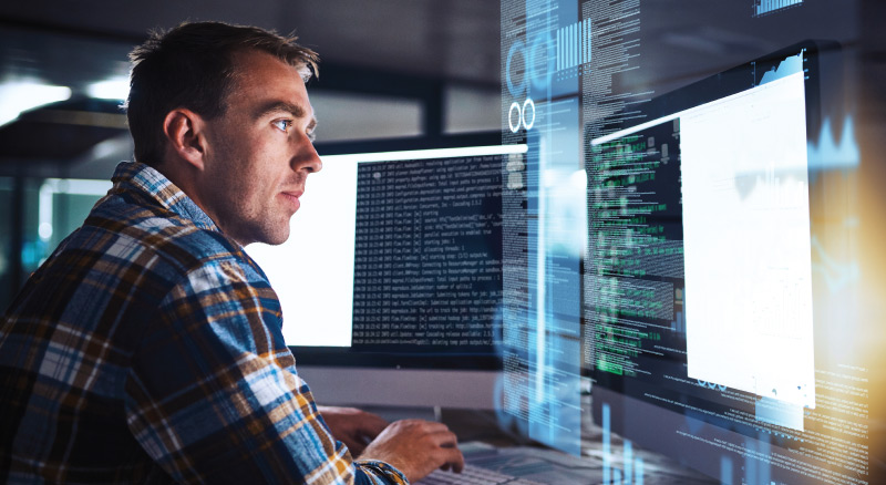 A person in a checkered shirt working on multiple computer screens displaying code and data visualizations