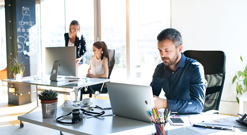 Three individuals working on laptops in a modern office space with large windows