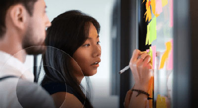 Two individuals engaged in a brainstorming session, writing on colorful sticky notes attached to a glass wall.