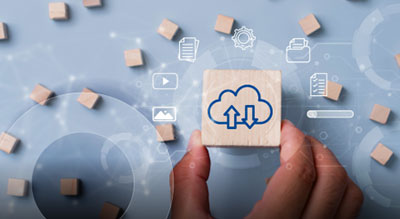 A hand holds a wooden cube with a cloud icon showing upload and download arrows. Several other wooden cubes with digital and cloud‑related symbols are scattered on a blue, tech‑themed background