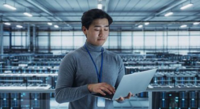 Worker holding a laptop, standing in a large server room filled with rows of server racks