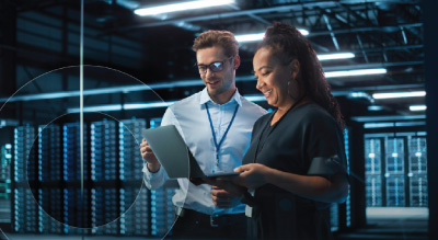 Two individuals in a server room examining a laptop, surrounded by servers and networking equipment.