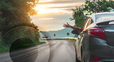 Car driving on a winding road with a person’s arm extended out of the window, surrounded by lush green trees and hills under a bright sunset sky