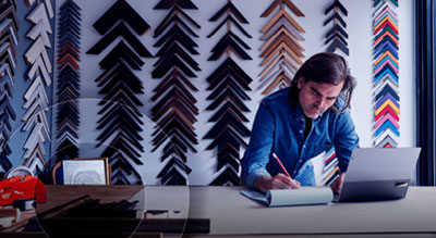 An individual is seated at a desk, writing in a notebook and working on a laptop, with colorful frames on the wall behind them