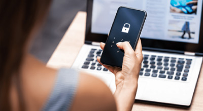 An individual sitting at a desk with an open laptop, holding a black phone displaying a white lock icon