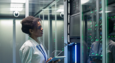 A person is standing in front of a server rack in a data center, inspecting the equipment with a tablet
