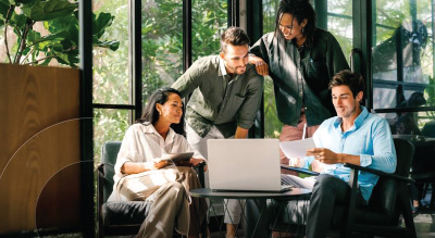 Four people are gathered around a laptop in a modern office, discussing documents and working together.