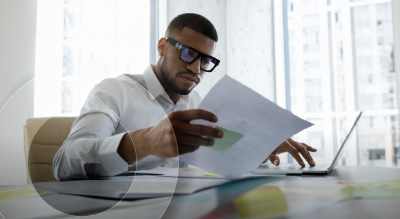 Man in glasses works intently, reviewing documents at a desk with a laptop