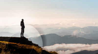A silhouette of a person standing on a mountain peak, overlooking a vast landscape of lower mountain ranges enveloped in clouds