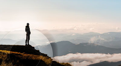 A person stands on a mountain peak, overlooking a vast landscape of clouds and mountains, illuminated by the sun’s rays