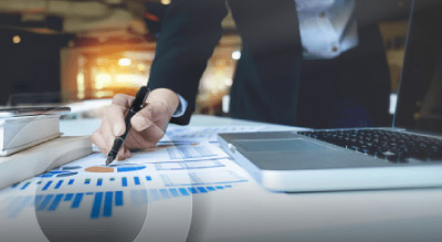 A person’s hand holding a pen over a chart on a desk with a laptop