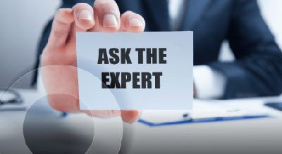 A business card with the text “Ask the Expert” is held by a hand in front of a blurry office scene
